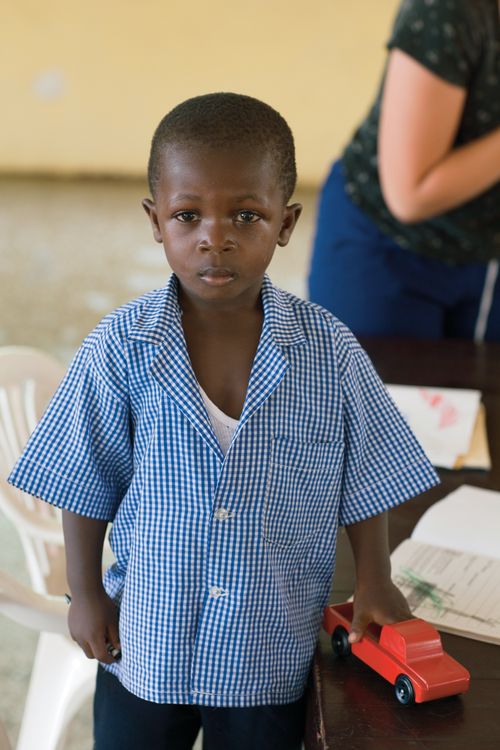 A little boy from Africa stands in a blue button-up shirt and holds a red toy truck in his hand.
