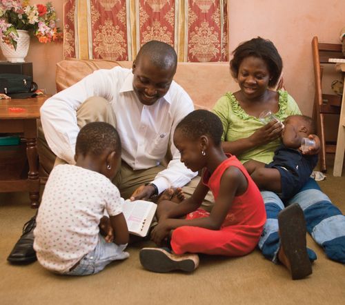 An African family seated in front of a sofa in their living room.  They are reading the scriptures.  Taken in Ghana, West Africa.