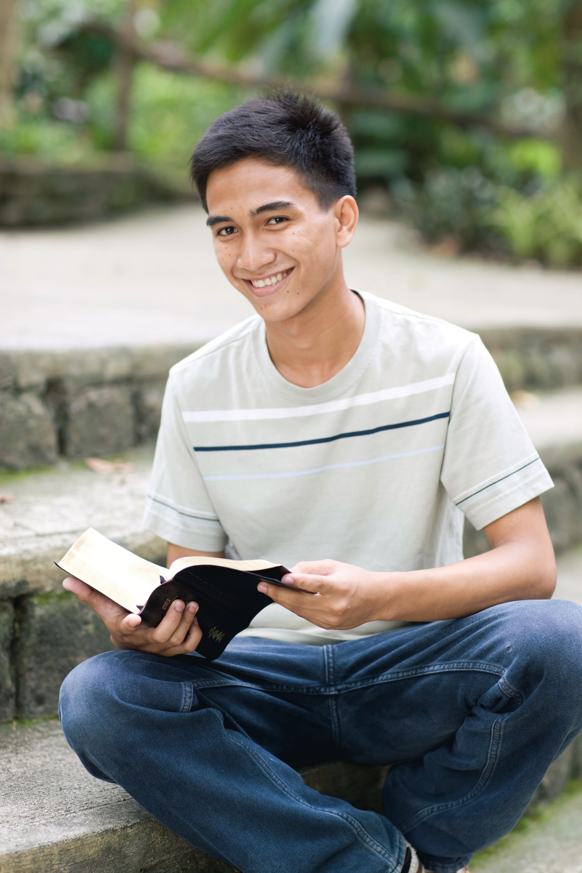 A young man sits outside on steps, with his scriptures open in his hands.