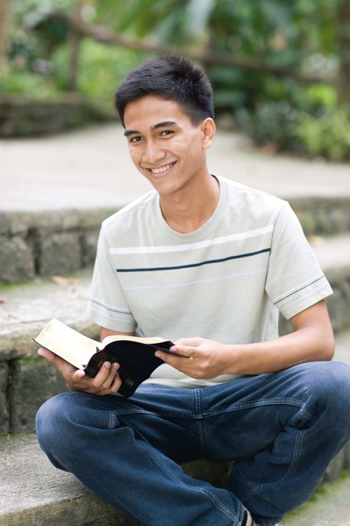 A young man sits outside on steps, holding an open set of scriptures and smiling.