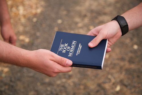 A hand is shown giving a blue paperback Book of Mormon to someone else.