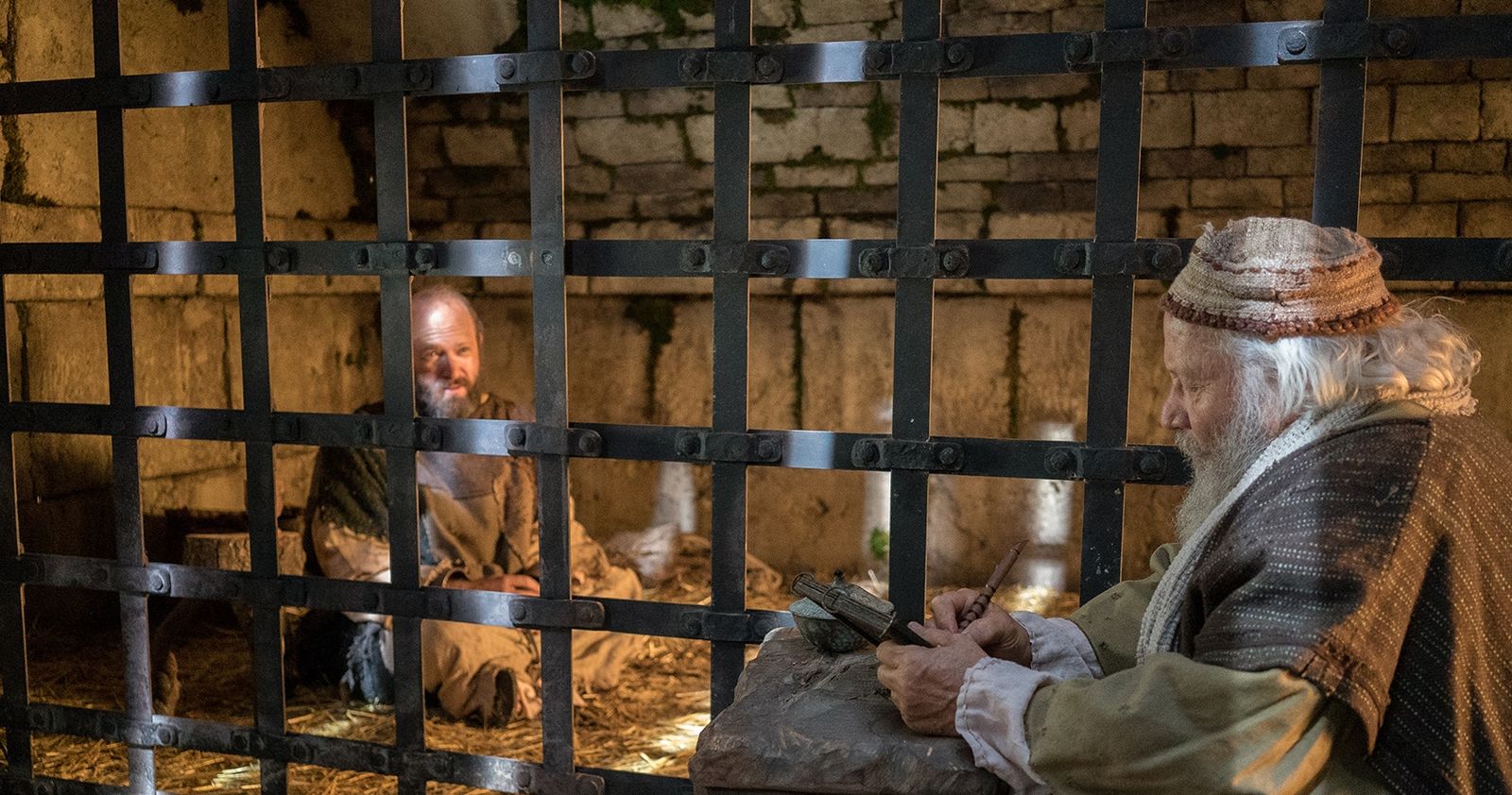 Paul, sitting in a prison cell with his hands shackled, dictates a letter to a scribe.