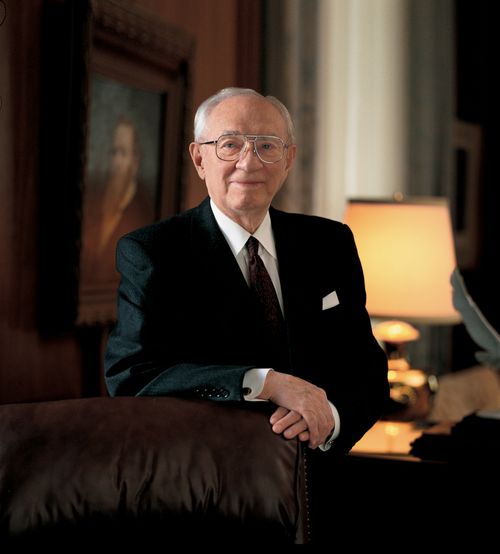 A portrait photograph by Jed A. Clark of Gordon B. Hinckley in a dark suit, standing with his arm resting on a leather chair.