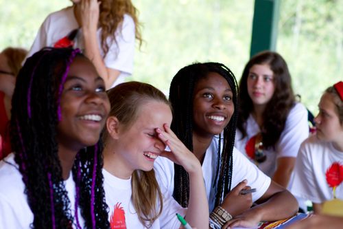 A group of girls in white T-shirts sit at a table outside and laugh during girls’ camp.