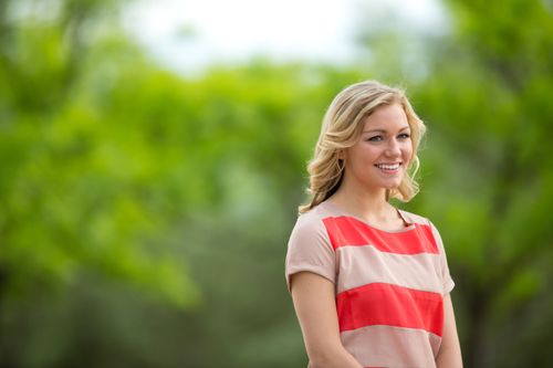 A portrait of a young woman with blonde hair and wearing a red and tan striped blouse, standing outside, looking to the side and smiling.