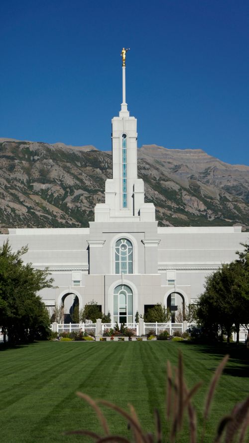 The front and spire of the Mount Timpanogos Utah Temple, with a green lawn leading to the temple and a mountain in the background.