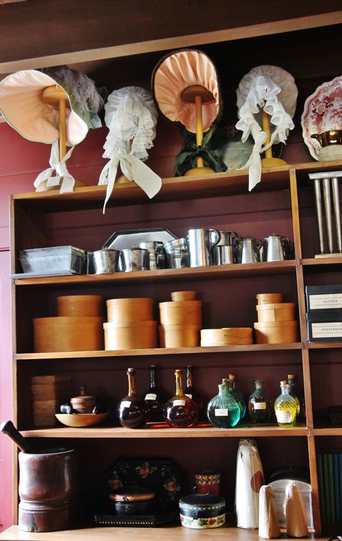 Shelves stacked with goods for purchase, such as round boxes, tin cups, glass bottles, and bonnets with ribbons.