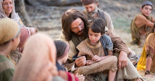 An actor portraying Jesus Christ holding a child on his lap. They are surrounded by people.