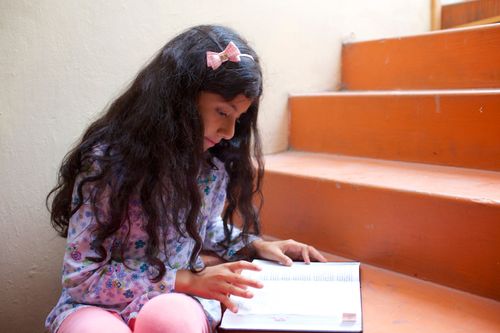 A young girl with a bow in her hair sits on the steps and reads from her scriptures.
