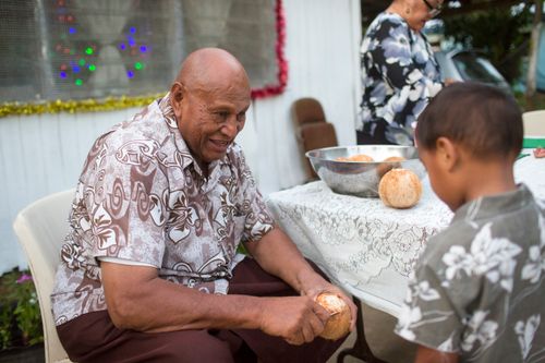 A man sits outside cutting a coconut and interacting with his young grandson.