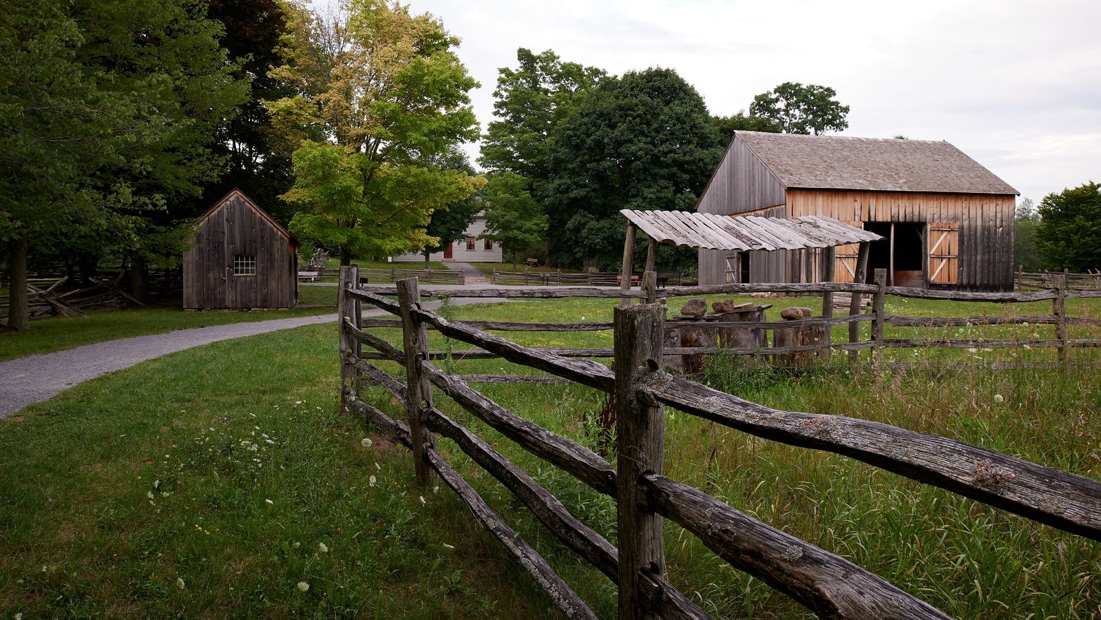 An exterior view of Joseph Smith's family exterior buildings in Palmyra, New York. 
There is farmland and a few smaller buildings on the land. They appear to be a barn and some kind of shed, likely another small barn or storage for a wagon.