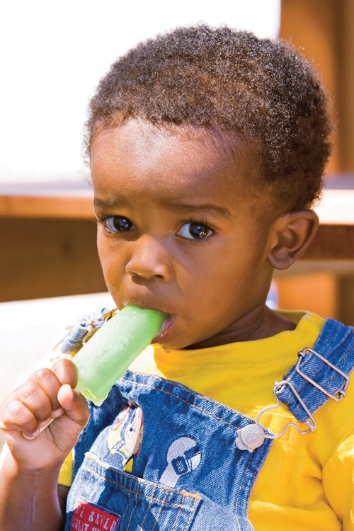 A toddler boy wearing overalls, licking a green Popsicle.