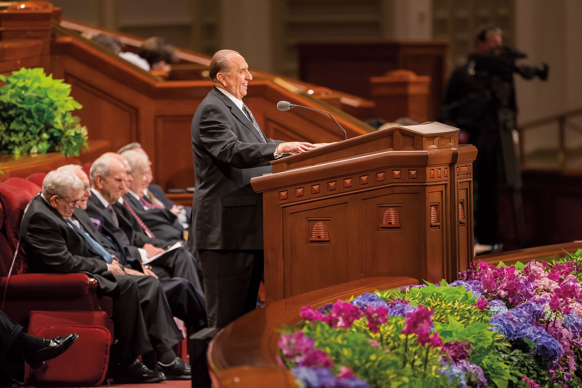 Thomas S. Monson standing at the pulpit at general conference, with several Apostles sitting behind him.