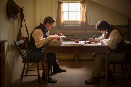 fotografia da dramatização de um filme, Joseph e Oliver sentados à mesa, frente a frente, Oliver escrevendo