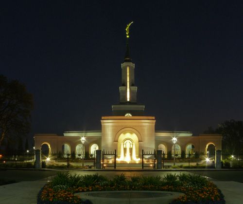 The front of the Sacramento California Temple lit up at night, with a view of the grounds, including the fountain out front, flowers, trees, and the angel Moroni on the spire.