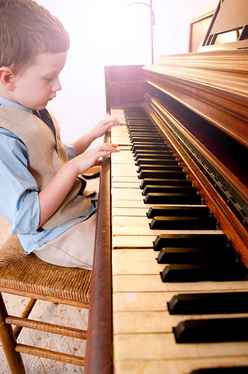 A little boy in a vest sits on a wooden chair and plays on a piano.