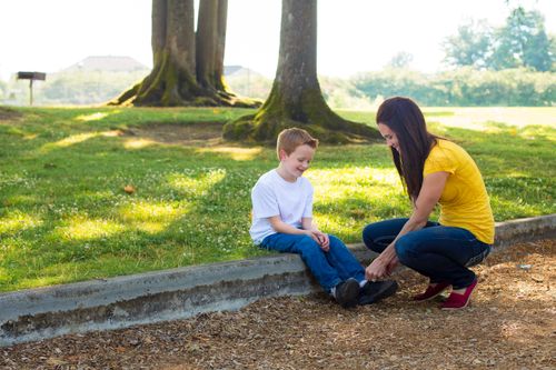 A mother helps tie her son’s shoes while outside at a park.