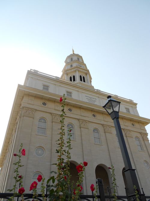 A view of the front of the Nauvoo Illinois Temple from the ground, looking up past pink flowers to the spire on the temple.