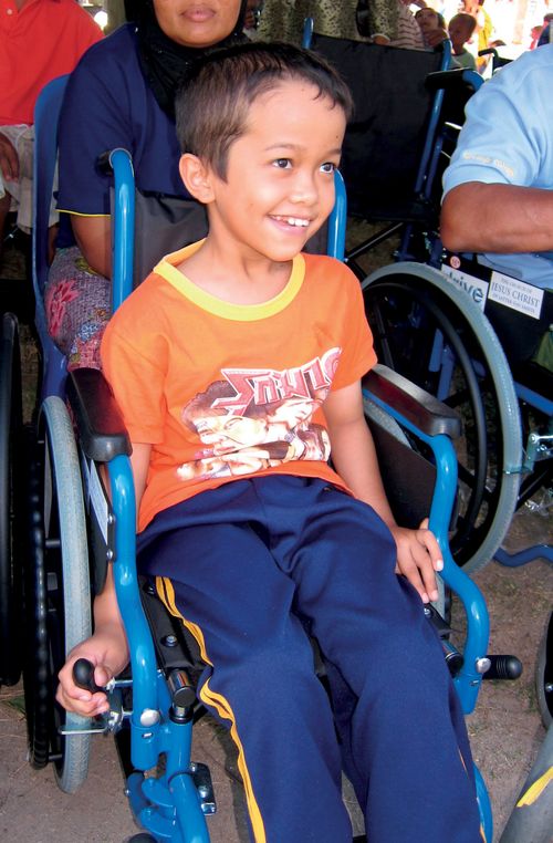 A young boy from Thailand in blue sweatpants and an orange shirt, smiling and sitting in a blue wheelchair.