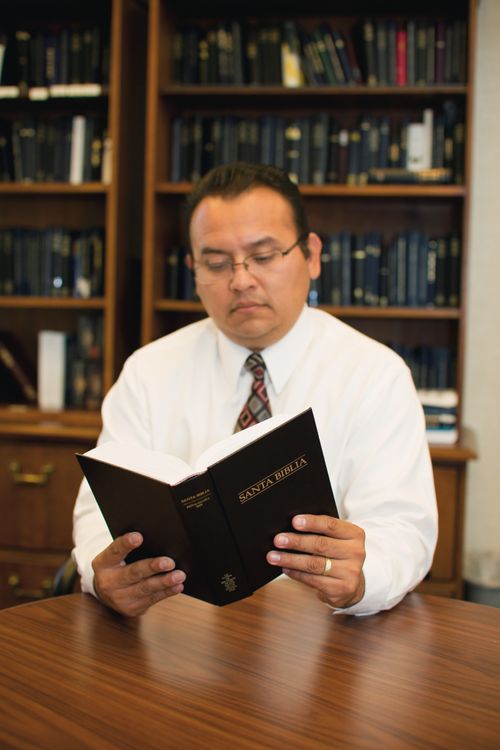 A man in a white shirt and tie sits at a table and reads a Spanish Bible, with several shelves of books seen in the background.