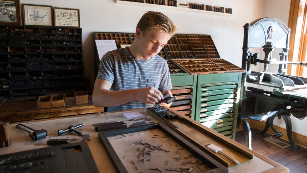 A young man uses historical tools to set type inside a print shop.