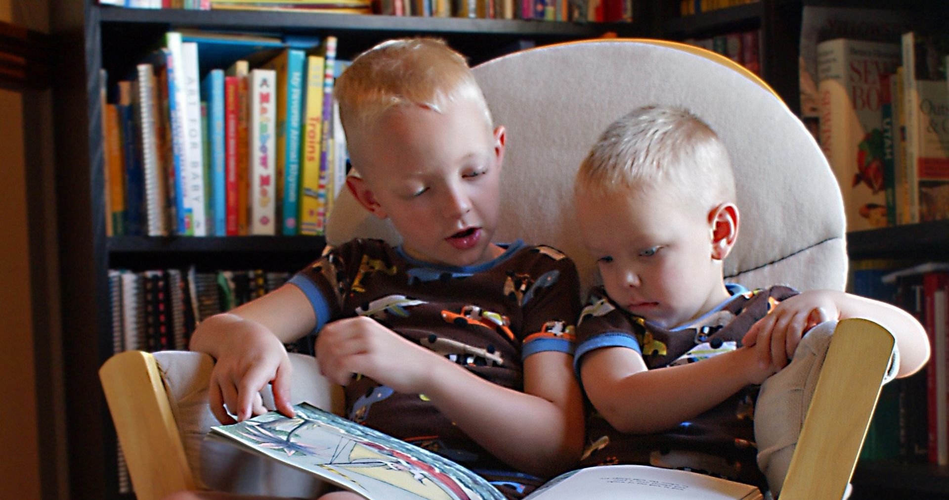 Two young brothers sit on a chair together. The older brother is reading a book to his younger brother.