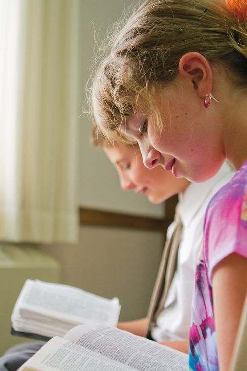 A girl sits next to a boy, and both have a set of scriptures open on their laps as they read from them while sitting in a classroom.