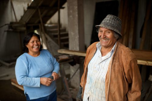 A man in a button-up shirt, brown jacket, and hat laughing and standing with his wife nearby in a long-sleeve blue shirt in a workshop in Ecuador.