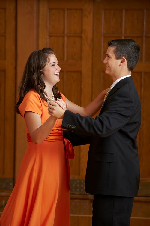 A young woman in a long orange dress dances with a young man in a suit.