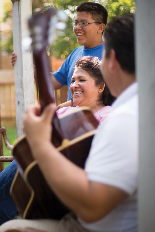 A young man is seen playing a guitar while his mother and brother stand nearby, singing.
