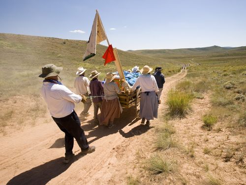 A man dressed as a pioneer pulls a rope attached to a handcart as it goes downhill, with a group of other men and women helping push the handcart.