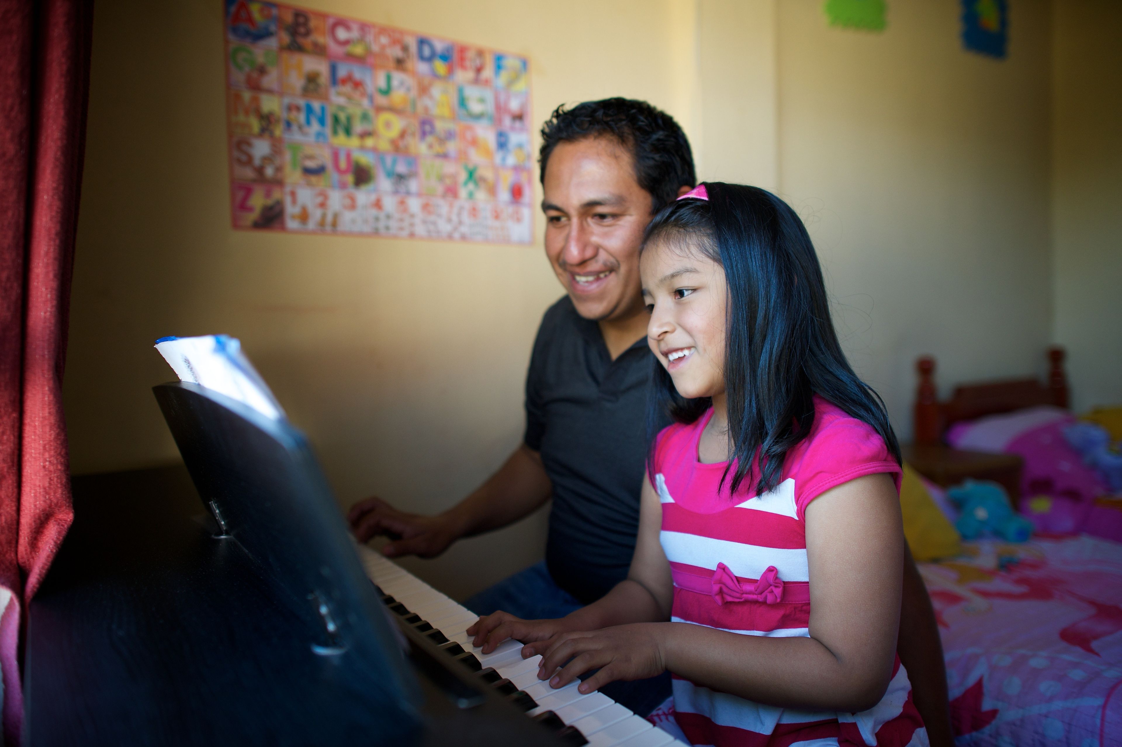 A father sits next to his daughter at a piano while they play together.