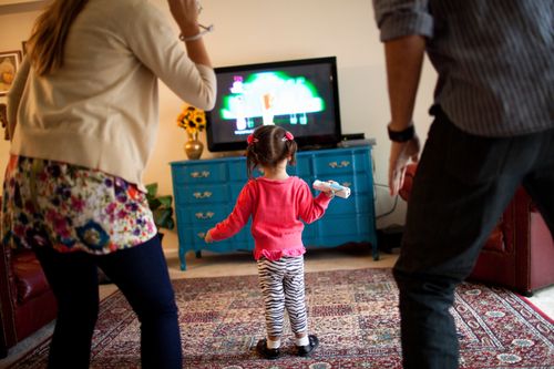 A mother and father standing while playing a video game on the TV with their young daughter standing in front of them in their living room.