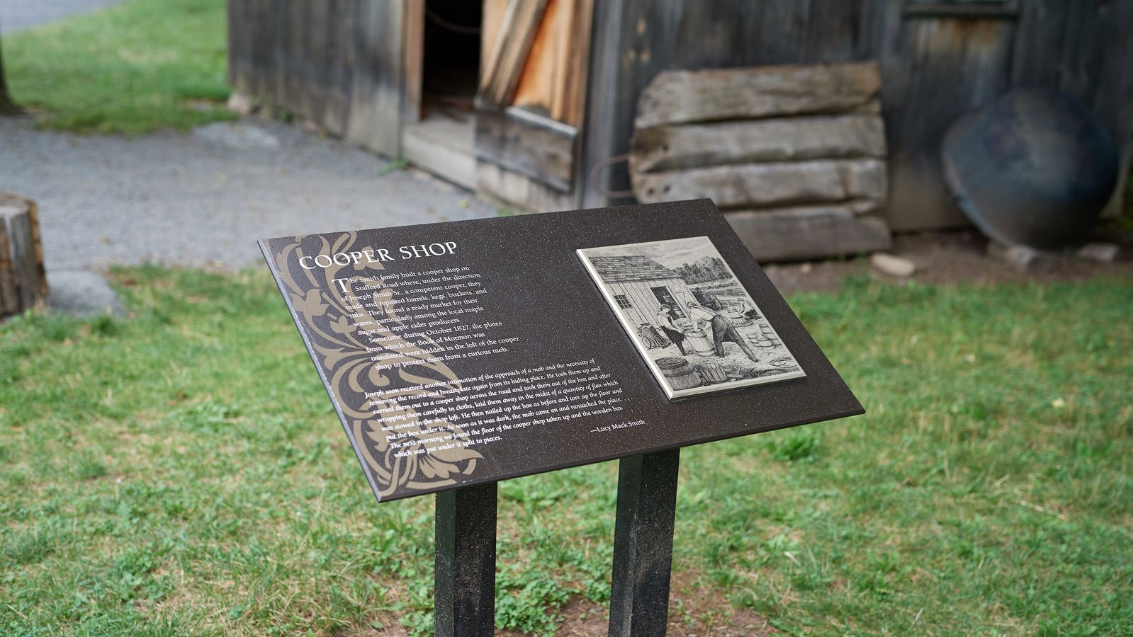 Various plaques around Palmyra and the Sacred Grove. This plaque says, 'Cooper Shop.' It is surrounded by farmland and a rough wooden building in the background.