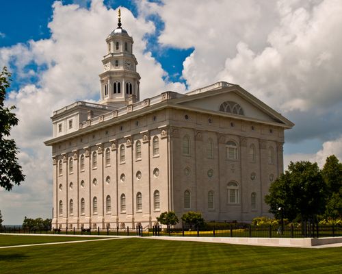 The back and side of the Nauvoo Illinois Temple, with a blue sky seen through the clouds and well-manicured green lawns in the foreground.