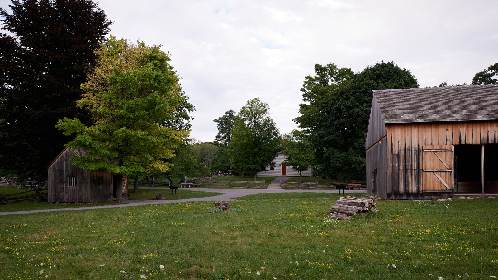 An exterior view of Joseph Smith's family exterior buildings in Palmyra, New York. 
There is farmland and a few smaller buildings on the land. They appear to be a barn and some kind of shed, likely another small barn or storage for a wagon.