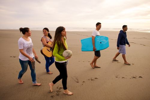 A group of youth on the beach, with one young woman carrying a ball, another carrying a guitar, a young man carrying a bodyboard, and another young man and young woman walking nearby.