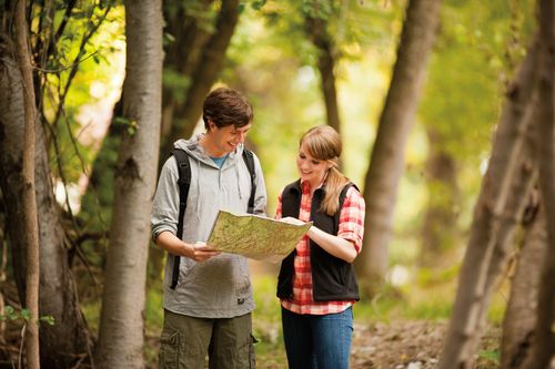 A young man holds a map and a young woman looks at it while they are on a hike in the woods.