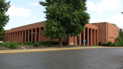 A large, colonnaded red brick building with an asphalt parking lot in the foreground.