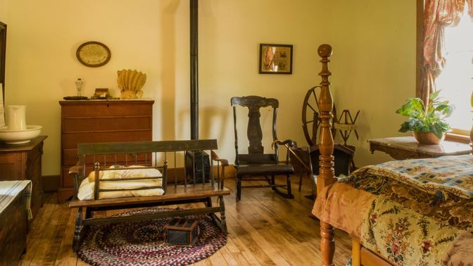 Interior room with yellow walls, rug, bed, pioneer sofa, rocking chair, wooden floor, and a cast iron stove.