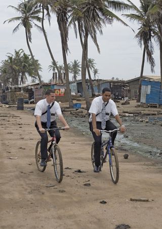 Caucasian and African elder missionaries riding their bikes down a dirt road through a town in Ghana, West Africa.