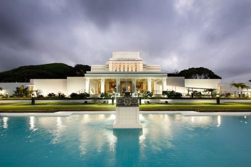 The front of the Laie Hawaii Temple at night, with the pool of water and fountain lit up near the entrance.