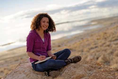 A young woman with brown curly hair, a purple shirt, jeans, and boots sits on a rock and writes in a journal.