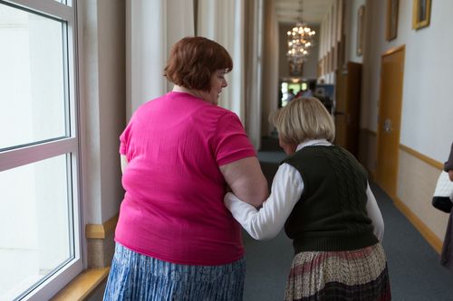 A woman in a pink shirt leading an older woman down the hallway of their meetinghouse.