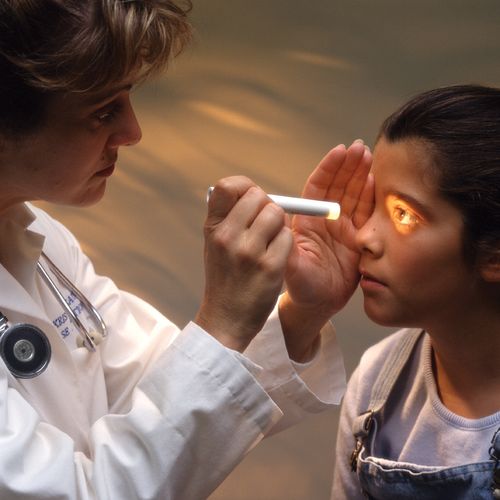 A female doctor in a white coat shining a flashlight in the eye of a young girl.