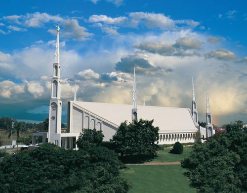 The Buenos Aires Argentina Temple seen from afar, with large green trees in the foreground and white clouds in the background.
