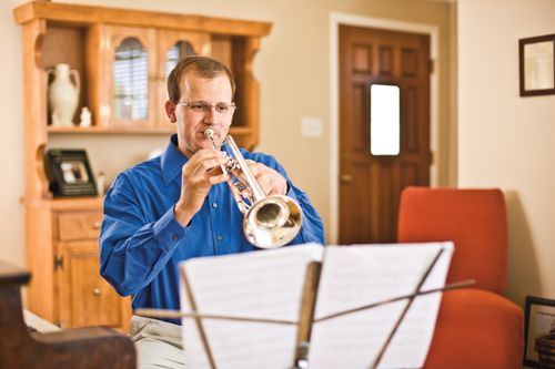 A man with glasses sits on a bench in the living room and plays his trumpet with music on a stand in front of him.