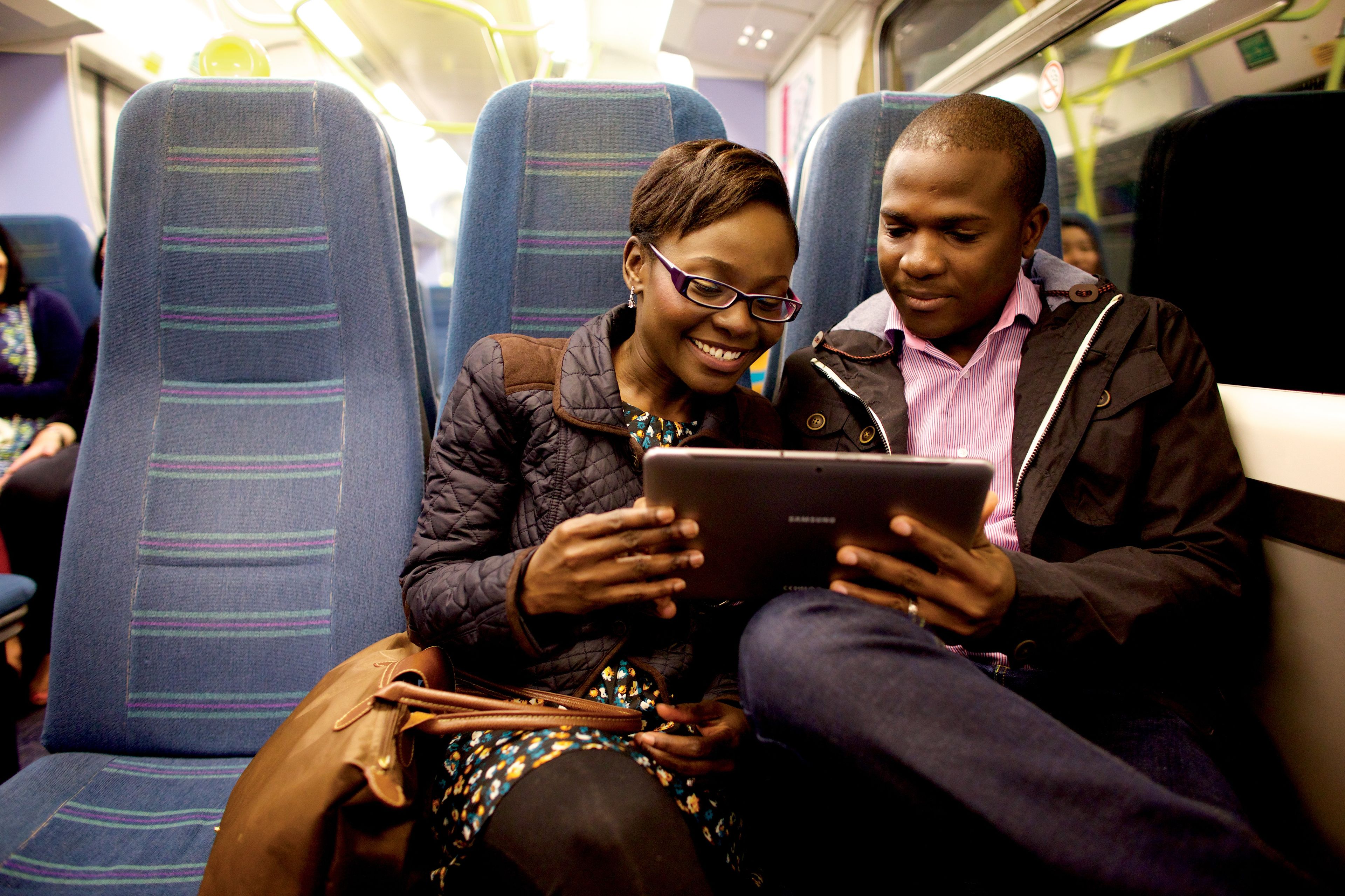A couple sitting on a train and looking at a tablet together.
