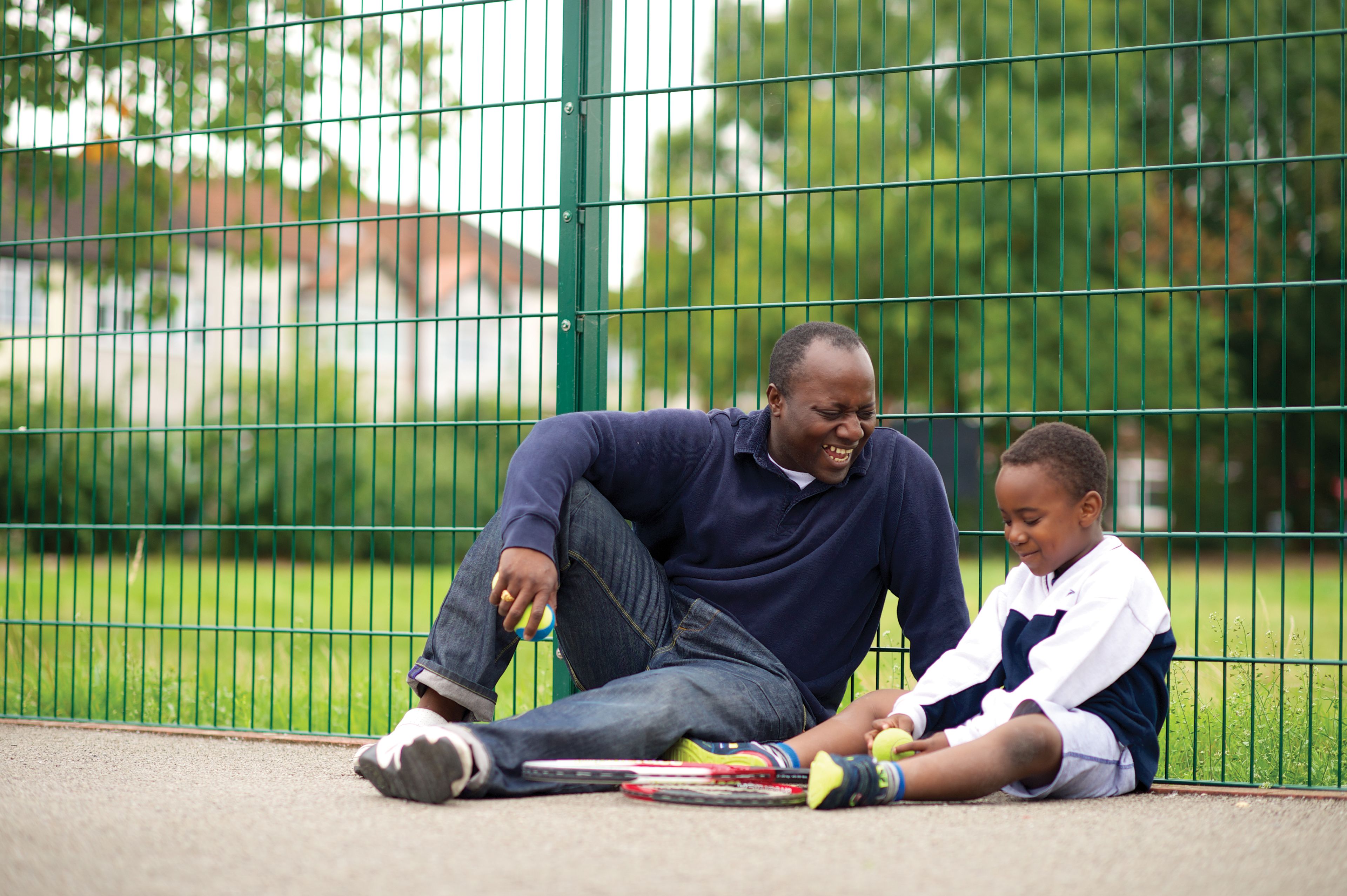 A father and son sitting on the ground in a tennis court.