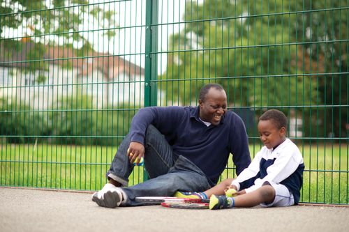 A father sits down next to his young son in a tennis court, with two tennis rackets beside them.